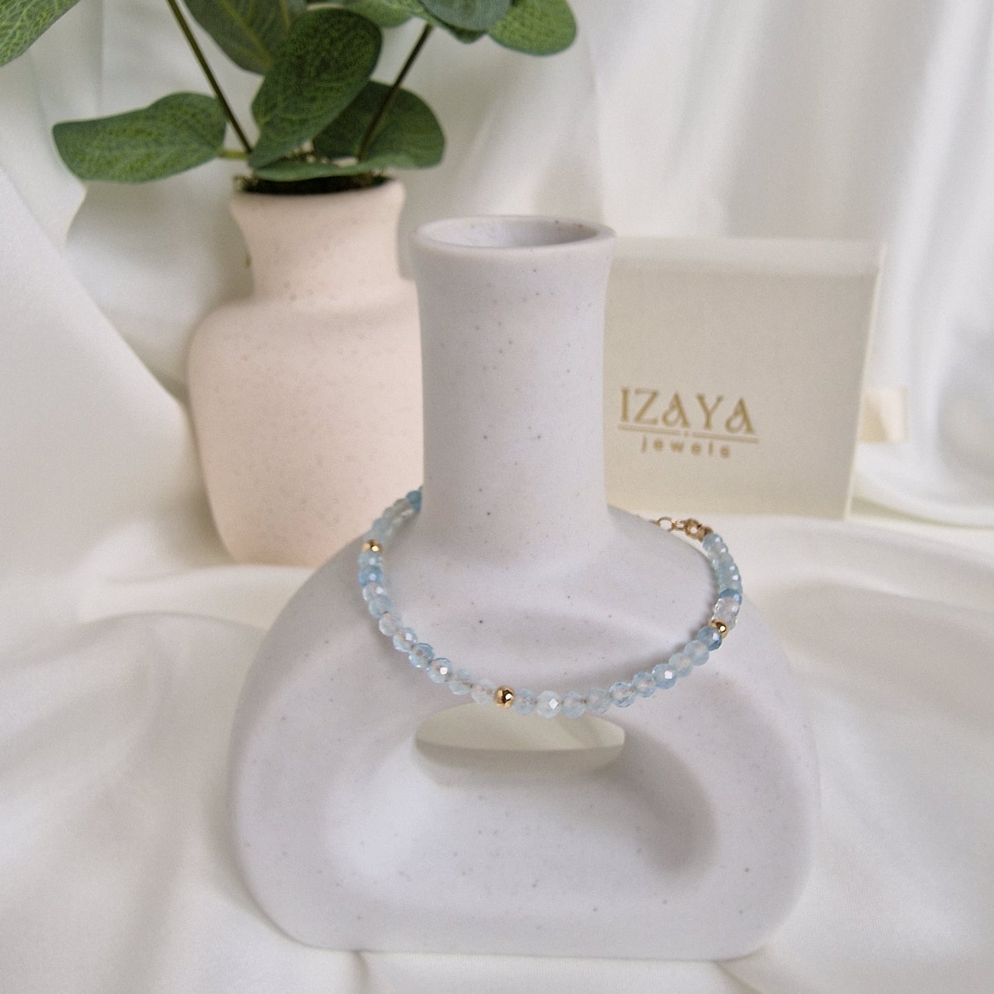 Blue beaded bracelet on a white mannequin arm with 'IZAYA' jewelry box in the background.