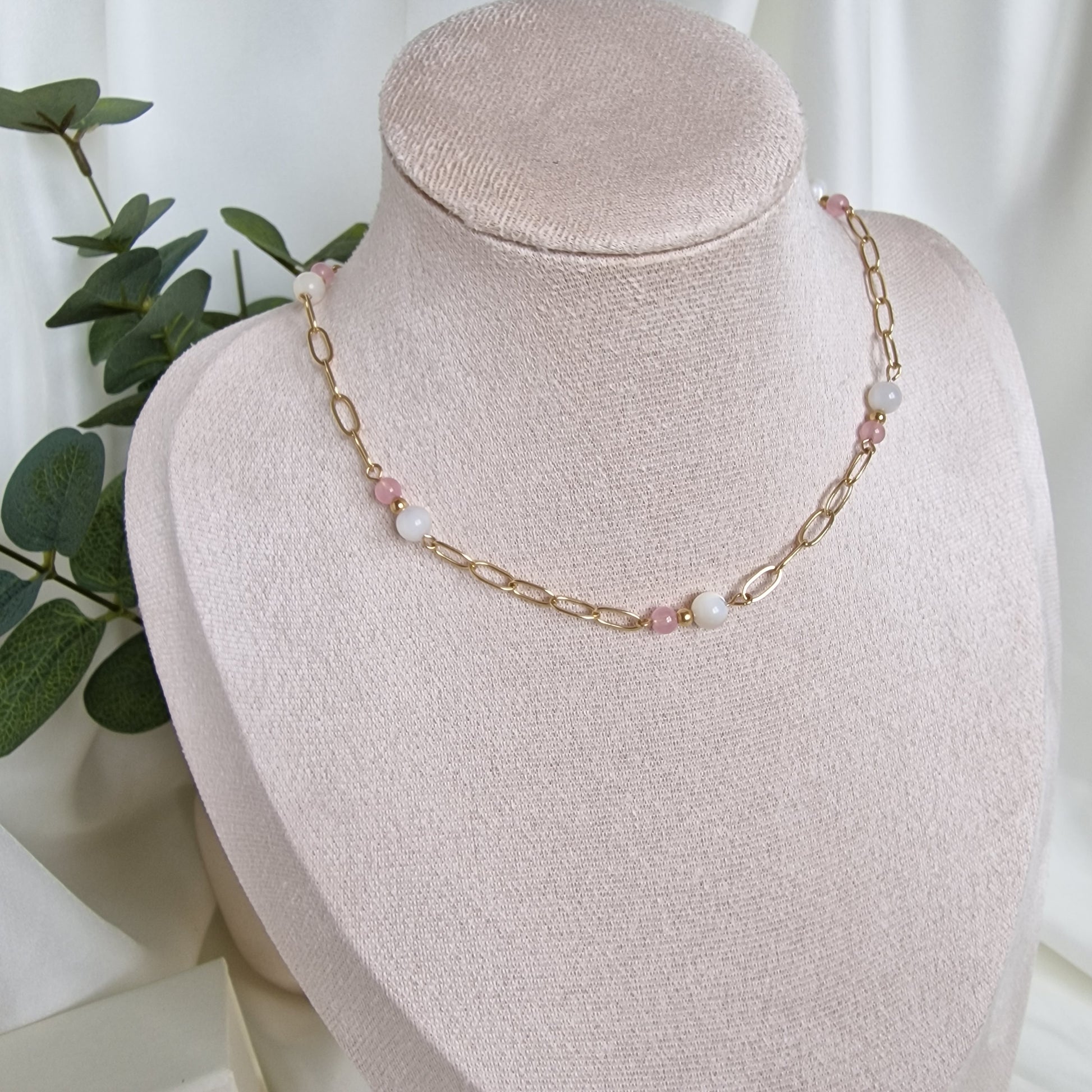 Gold-filled chain necklace with pink and white beads on a pink velvet stand with green leaves in the background